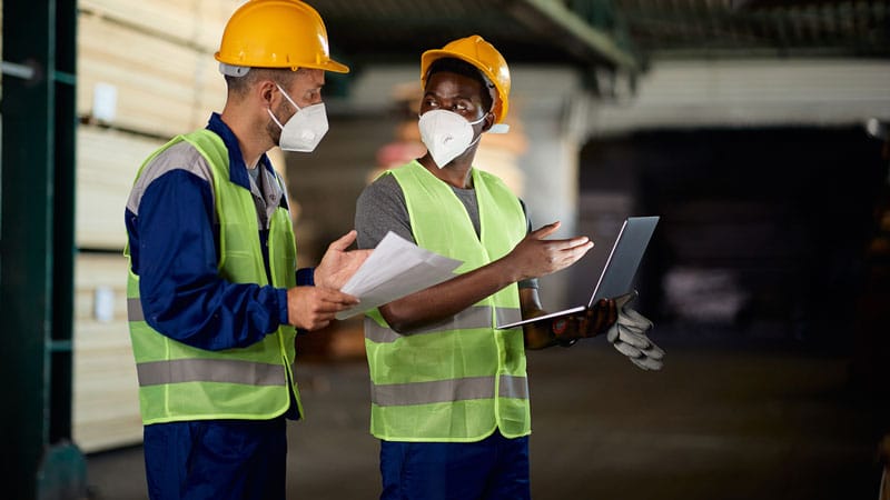 African American worker and his colleague using laptop and communicating while working at lumberyard warehouse